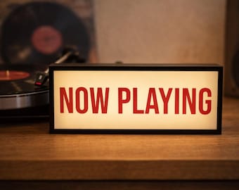 May include: A rectangular light box with a black frame and a white illuminated panel featuring the text NOW PLAYING in bold red capital letters. The sign is positioned horizontally on a wooden surface near a vinyl record player.