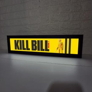 Puede incluir: Un letrero rectangular con marco negro con las palabras "KILL BILL" en letras amarillas grandes. La imagen presenta a una mujer con un mono amarillo sosteniendo una espada. Las palabras "VOLUME 2" son rojas. El fondo es amarillo.