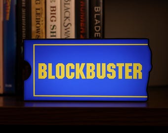以下が含まれることがあります： 映画レンタルカードを思わせる青と黄色のBlockbusterの看板が棚に置かれています。「BLOCKBUSTER」の文字が黄色で表示されています。背景には本が見えます。