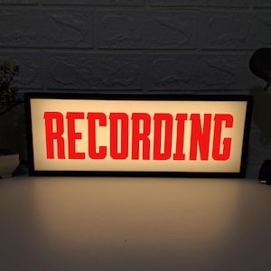 Può includere: Un cartello nero incorniciato con sfondo bianco e scritta rossa che dice "RECORDING".