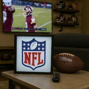 Op de afbeelding: Een ingelijst NFL-logo met een blauw schild, witte voetbal en sterren en rode letters. Een bruine Wilson-voetbal ligt naast het frame op een houten tafel. Een tv-scherm toont een American football-wedstrijd op de achtergrond.