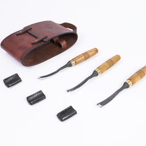 以下が含まれることがあります： 木製の柄と金属製の刃が付いた木彫り用のノミ3本セット、茶色の革製ツールロール、黒革製の刃保護カバー3つ。白い表面に配置されたツール。