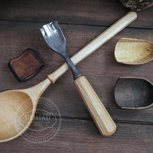 May include: Two wooden spoons and a wood carving tool with a leather sheath. The spoons are made of light wood and have a natural finish. The carving tool has a metal blade and a wooden handle. The tools are arranged on a wooden surface.