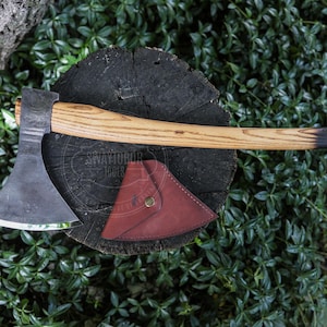 Könnte beinhalten: Eine handgeschmiedete Stahlaxt mit einem geschwungenen Holzstiel mit dunklem Ende, begleitet von einer dreieckigen, rotbraunen Lederscheide mit Messingdruckknopf. Der Axtkopf verfügt über eine breite, geschärfte Klinge und ein dunkles Finish.