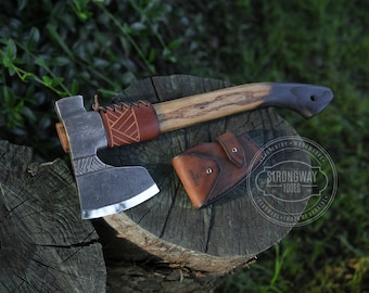 May include: Axe with a wooden handle and a leather sheath. The axe head is made of metal and has a sharp blade. The handle is a light brown color with a darker brown grip. The leather sheath is brown and has a button closure. The axe is resting on a tree stump.