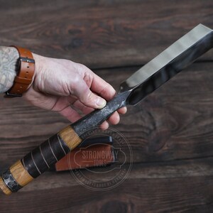 May include: A hand holds a long, thin, metal chisel with a wooden handle wrapped in black leather. The chisel has a beveled edge and a rounded end. The handle is secured with a metal band. The chisel is labeled "Strongway Tools" with the text "Handmade - Forge of Viking".