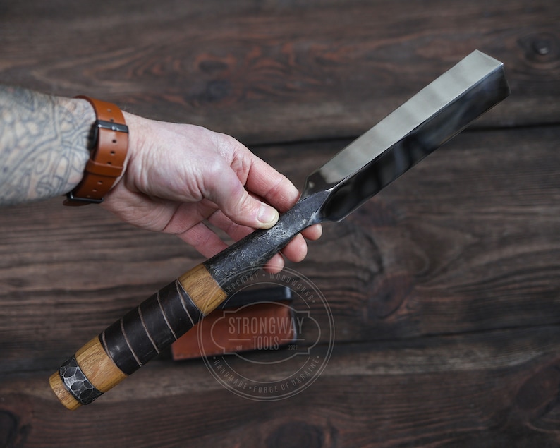 May include: A hand holds a long, thin, metal chisel with a wooden handle wrapped in black leather. The chisel has a beveled edge and a rounded end. The handle is secured with a metal band. The chisel is labeled "Strongway Tools" with the text "Handmade - Forge of Viking".