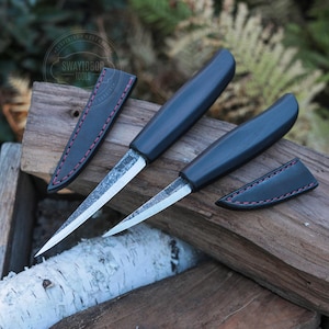 Puede incluir: Dos cuchillos con mangos oscuros, hojas plateadas y fundas de cuero negro con costuras rosas descansan sobre una superficie de madera. Los cuchillos están en ángulo, mostrando sus puntas afiladas. El logotipo "SWAYTOBOR TOOLS" es visible.