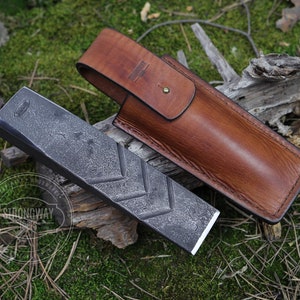 Puede incluir: Una cuña de metal negro con una superficie texturizada y una funda de cuero. La cuña mide aproximadamente 25 cm de largo y tiene una ligera curva. La funda es de cuero marrón con una costura cosida.
