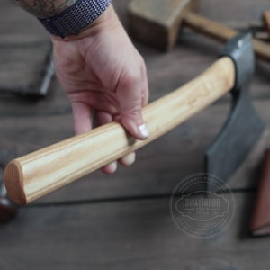 May include: A wooden-handled adze with a black metal blade. The adze is held in a hand, and the handle is made of light and dark wood.
