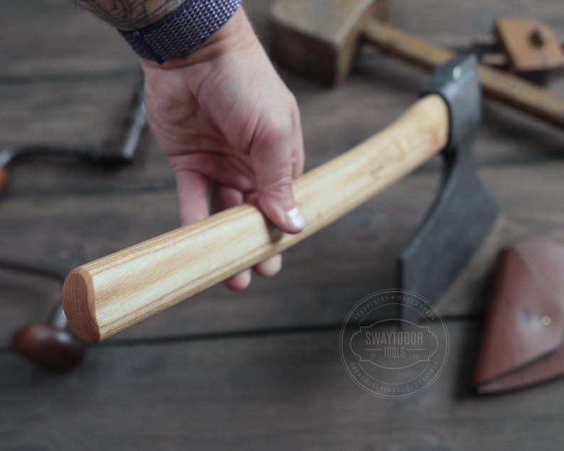 May include: A wooden-handled adze with a black metal blade. The adze is held in a hand, and the handle is made of light and dark wood.
