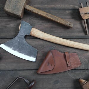 May include: A hand-forged axe with a wooden handle and a leather sheath. The axe has a curved blade and a sharp point. The sheath is brown leather with a decorative stitching detail. The axe is resting on a wooden surface.
