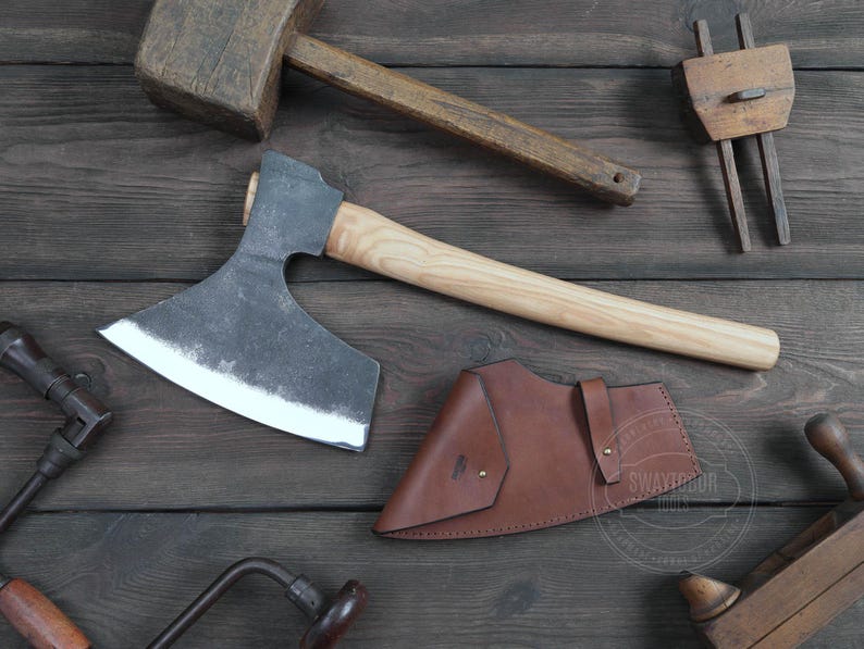 May include: A hand-forged axe with a wooden handle and a leather sheath. The axe has a curved blade and a sharp point. The sheath is brown leather with a decorative stitching detail. The axe is resting on a wooden surface.