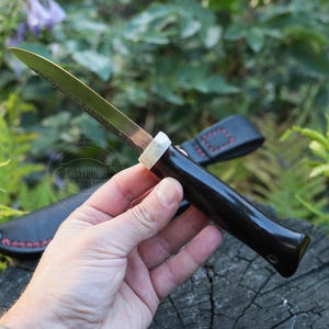 May include: A handcrafted knife with a dark wooden handle and a silver blade. The knife is held in a hand, with a black leather sheath visible in the background. The knife has a unique design and is likely used for cutting or crafting.