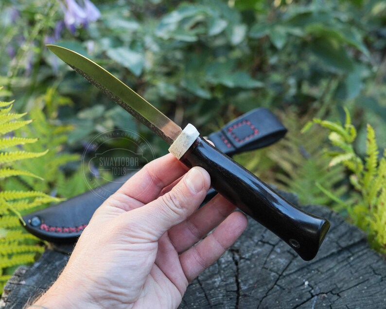 May include: A handcrafted knife with a dark wooden handle and a silver blade. The knife is held in a hand, with a black leather sheath visible in the background. The knife has a unique design and is likely used for cutting or crafting.