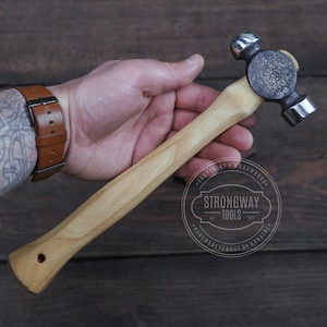 May include: A wooden-handled ball-peen hammer with a black metal head. The hammer is held in a hand wearing a brown leather wristwatch. The hammer is labelled "Strongway Tools" with the text "Handmade Forge of Ukraine".