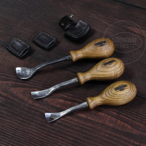 May include: Three wood carving tools with wooden handles and metal blades. The tools are on a dark wood surface. The handles are a light brown color and have a grain pattern. The blades are silver and have a slight curve. The tools are labeled "Strongway Tools".