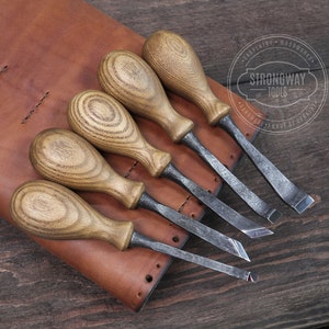 Op de afbeelding: Set van zes beitel met houten handvatten en een leren rolhoes. De beitels hebben verschillende vormen en zijn gerangschikt in de hoes. De hoes heeft de tekst "Strongway Tools" erop.