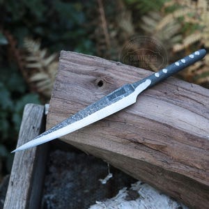 May include: A handmade knife with a silver blade and a dark gray handle with white dot accents. The knife is resting on a weathered wooden surface, showcasing its sharp design and craftsmanship. The blade is approximately 8 inches long.