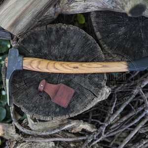Könnte beinhalten: Ein Handwerkzeug mit einem dunklen Metallkopf und einem geschwungenen Holzgriff mit schwarz gebeiztem Ende. Unter dem Werkzeug befindet sich eine kleine, rotbraune Lederscheide mit einem Metalldruckknopf. Der Text SWAYTOBOR TOOLS ist sichtbar.