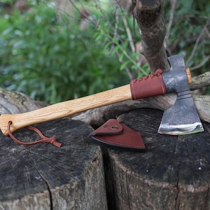 Peut inclure: Une hachette avec un manche en bois clair, une tête en métal forgé sombre et une gaine en cuir marron fixée par un laçage. Un étui en cuir marron avec un bouton-pression en métal est posé à côté, et un cordon en cuir est fixé à la base.
