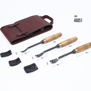 Może przedstawiać: Zestaw trzech narzędzi do rzeźbienia w drewnie z drewnianymi uchwytami i metalowymi ostrzami, wraz z brązowym sk&oacute;rzanym etui i trzema czarnymi sk&oacute;rzanymi osłonami ostrzy. Narzędzia mają 12 cm długości.