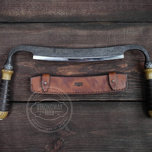 Op de afbeelding: Een gebogen, handgesmeed houtbewerkingsgereedschap met een leren schede. Het gereedschap heeft een houten handvat en een metalen blad. De schede is van bruin leer met stiksels en een metalen klinknagel. Het gereedschap ligt op een houten oppervlak. De tekst "Strongway Tools" is zichtbaar op de schede.