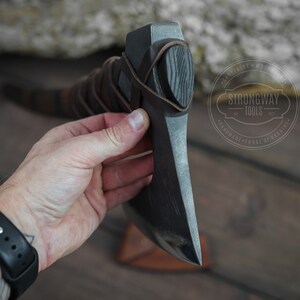 May include: A black axe with a leather handle and a wooden grip. The axe is held in a hand and the background is a wooden surface. The axe has a Strongway Tools logo on it.