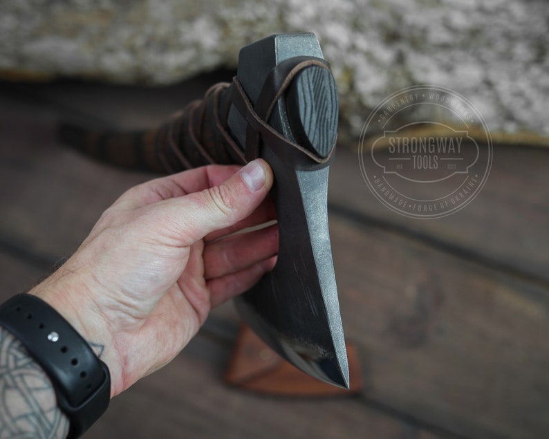 May include: A black axe with a leather handle and a wooden grip. The axe is held in a hand and the background is a wooden surface. The axe has a Strongway Tools logo on it.