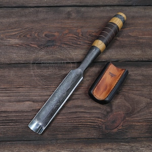 May include: A long, thin, metal chisel with a wooden handle and a leather sheath. The chisel has a black and brown handle with a striped pattern. The sheath is black with a brown leather flap and the text "Strongway Tools" printed on it.