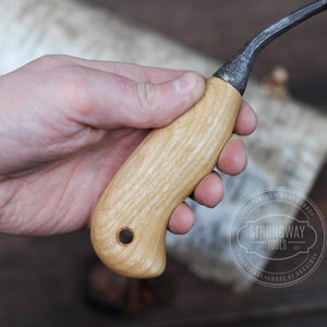 May include: A wooden handled carving knife with a curved blade. The handle is made of light wood with a grain pattern. The blade is made of dark metal and has a slight curve. The knife is held in a hand.