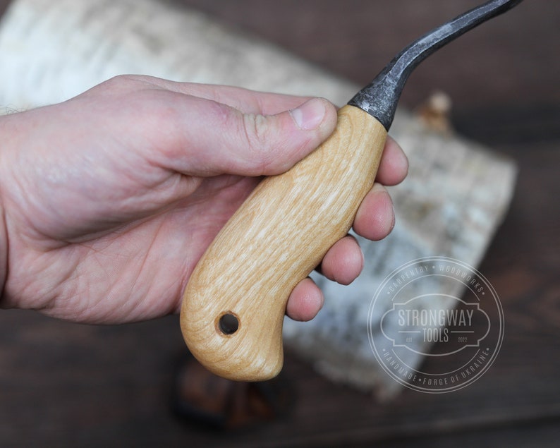 May include: A wooden handled carving knife with a curved blade. The handle is made of light wood with a grain pattern. The blade is made of dark metal and has a slight curve. The knife is held in a hand.