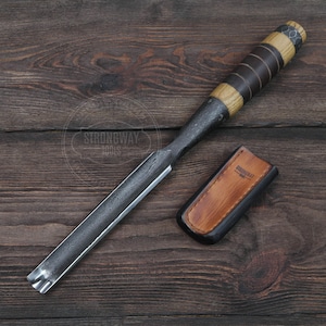 May include: A wood carving chisel with a wooden handle and a leather sheath. The chisel is made of metal and has a sharp blade. The sheath is brown leather with the word "Strongway Tools" embossed on it.