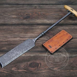 May include: A hand-forged metal tool with a wooden handle and a leather sheath. The tool has a long, flat blade and a T-shaped handle. The sheath is brown and has a stitched border. The tool and sheath are on a wooden surface.