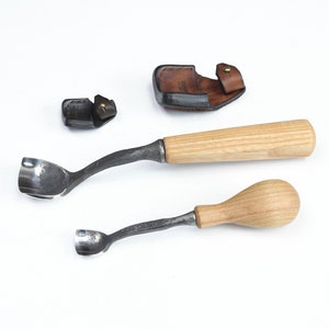 Pu&ograve; includere: Due strumenti per intagliare il legno con manici in legno e lame in metallo. Gli strumenti sono su una superficie bianca. &Egrave; mostrato anche un fodero in pelle marrone.