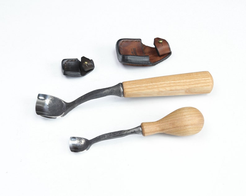 Pu&ograve; includere: Due strumenti per intagliare il legno con manici in legno e lame in metallo. Gli strumenti sono su una superficie bianca. &Egrave; mostrato anche un fodero in pelle marrone.