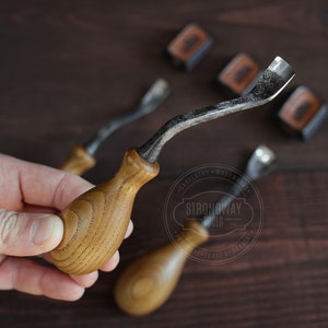 May include: Three wood carving tools with curved blades and wooden handles. The tools are on a dark wood surface. The tools are labeled "Strongway Tools".