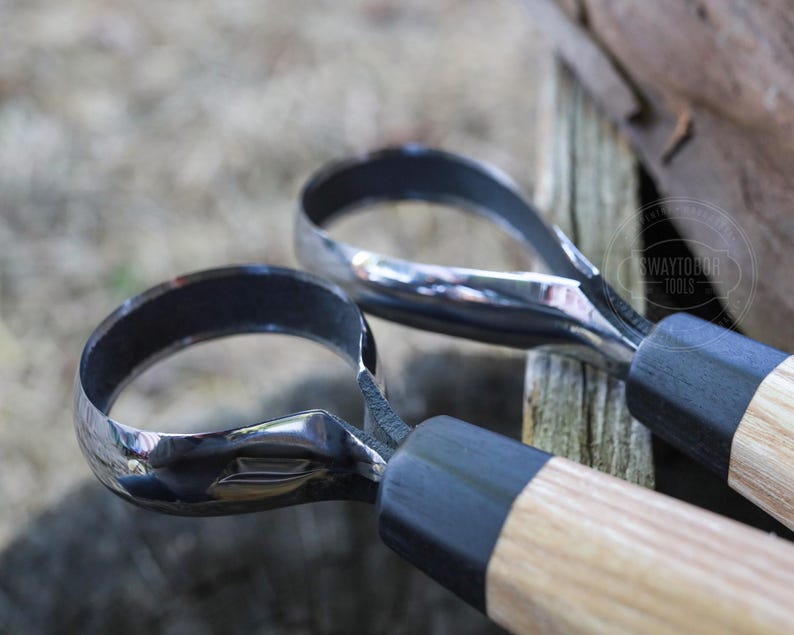 May include: Close-up of two metal carving tools with curved blades and wooden handles. The blades are shiny and reflect the light, while the handles have a natural wood grain. The tools are designed for woodworking.