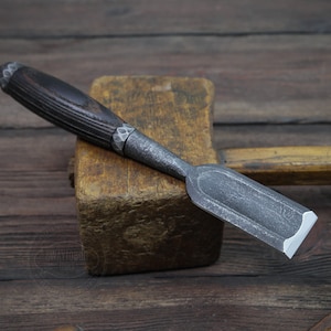 Könnte beinhalten: Nahaufnahme eines Holzmeißels mit einem dunklen Holzgriff und einer Metallklinge. Der Meißel liegt auf einem Holzblock.