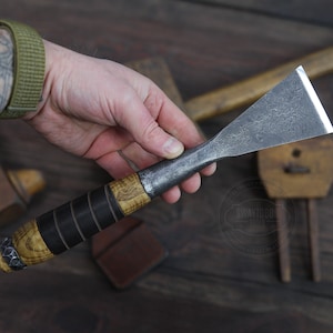 以下が含まれることがあります： 手は、幅広で平らな刃を持つ、木製の柄の金属製のノミを持っています。ノミは、柄の周りに黒い革の巻き付けがあります。背景は木製の表面です。