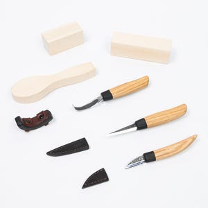 May include: A selection of wood carving tools and accessories, including three knives with wooden handles and black accents, two wooden blocks, a spoon-shaped wood piece, and a leather sheath. The tools are arranged on a white surface.
