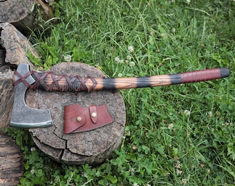 Può includere: Un'ascia in metallo con lama incisa e manico in legno avvolto da lacci in pelle marrone e un'impugnatura rosso scuro. Un fodero in pelle marrone abbinato con due bottoni a pressione in metallo color ottone è posizionato vicino alla testa.