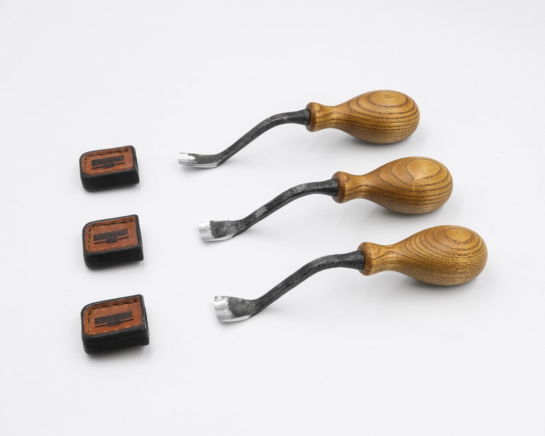 May include: Three wood carving tools with wooden handles and metal blades. The tools are on a white surface.