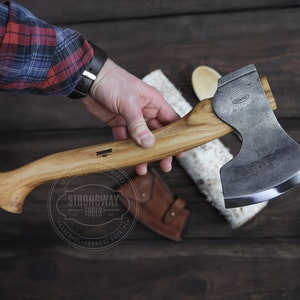Puede incluir: Una mano sostiene un hacha con mango de madera y cabeza de metal negro. El hacha tiene una hoja ligeramente curvada y una funda de cuero. El hacha está sobre una superficie de madera.