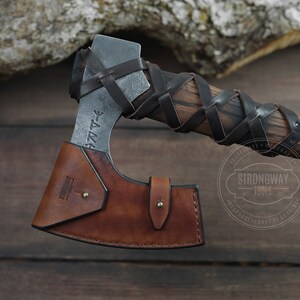 May include: A brown leather axe sheath with a black leather strap and a metal axe head with a wooden handle wrapped in black leather. The axe head has runes engraved on it and the text "Strongway Tools" is stamped on the sheath.
