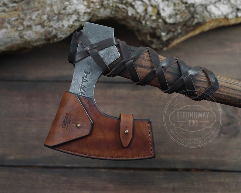 May include: A brown leather axe sheath with a black leather strap and a metal axe head with a wooden handle wrapped in black leather. The axe head has runes engraved on it and the text "Strongway Tools" is stamped on the sheath.