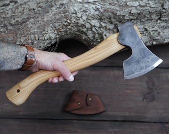 May include: A hand holds a small, single-bevel axe with a wooden handle. The axe has a dark gray finish and a sharp blade. The handle is made of light wood and has a slight curve. The axe is resting on a wooden surface.
