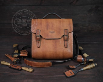 May include: A brown leather tool bag with a shoulder strap and buckles. The bag is filled with various woodworking tools, including a small axe, chisels, and a plane. The bag has a label that reads "Strongway Tools" and "Handmade Forged by Hand".