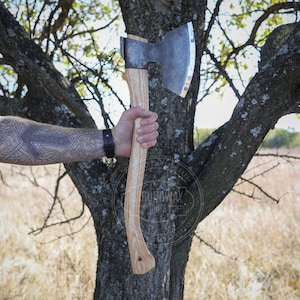 Puede incluir: Una persona sosteniendo un hacha grande con un mango de madera. El hacha tiene una hoja curva con un borde plateado. La persona tiene un tatuaje en el brazo.