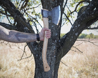 May include: A person holding a large,  axe with a wooden handle. The axe has a curved blade with a silver edge. The person is wearing a tattoo on their arm.